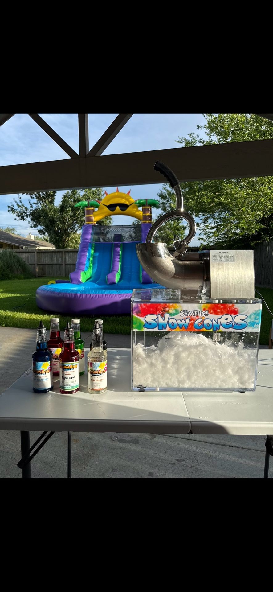 Snow cone machine and flavored syrups on a table with an inflatable water slide in the background.